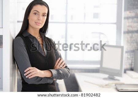 Confident office worker woman standing at desk, smiling at camera with arms folded.?