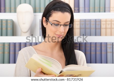 Smiling woman wearing glasses reading book in front of book shelf.?