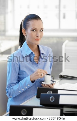 Smiling office worker girl having coffee, sitting at desk, looking at camera.?