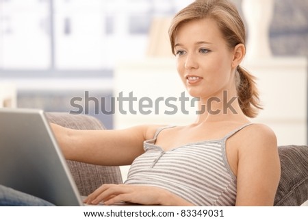 Casual young woman lying on sofa at home, browsing internet on laptop. Looking at screen, smiling.?