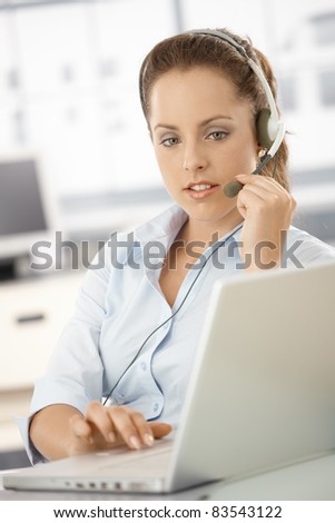 Attractive customer servicer working in bright office, using headphones and laptop.?