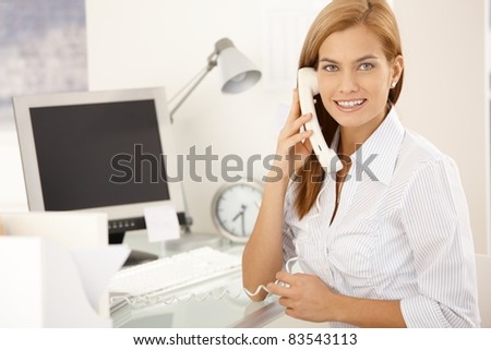 Happy office girl on landline phone call, sitting at desk, smiling at camera.?