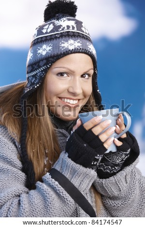 Attractive young female dressed up warm for skiing wearing cap and gloves drinking hot tea smiling front of winter landscape .?