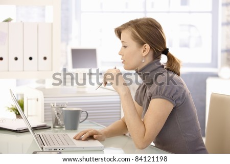 Attractive office worker sitting at desk, using laptop computer, looking at screen. Side view.?