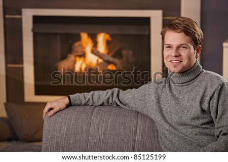Young man sitting on sofa at home on a cold winter day in front of fireplace, smiling.?