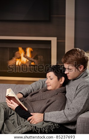 Young couple hugging on sofa in front of fireplace at home, reading book.?