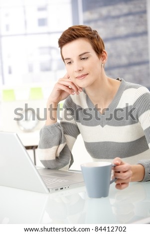 Young woman sitting at table, reading on laptop computer with coffee handheld, smiling?