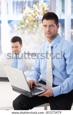 Handsome businessman sitting on top of desk, working on laptop.?