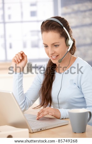 Young attractive customer servicer talking on headphones, using laptop, smiling.?