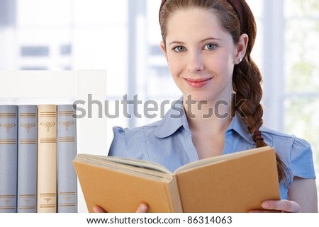 Portrait of attractive young woman standing in living room at home, reading a book, smiling.?
