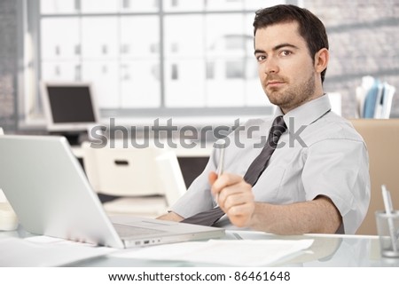 Young man working in bright office, sitting at desk, using laptop.?