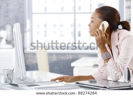 Busy office girl looking at computer screen while on landline phone call bending over office desk.?