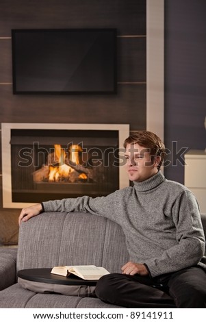 Young man sitting on sofa at home on a cold winter day, reading book in front of fireplace.?