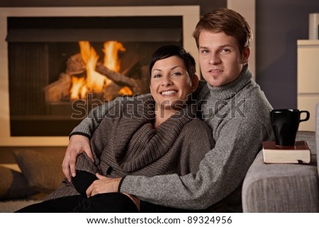 Young couple sitting on floor hugging in front of fireplace at home, looking at camera, smiling.?