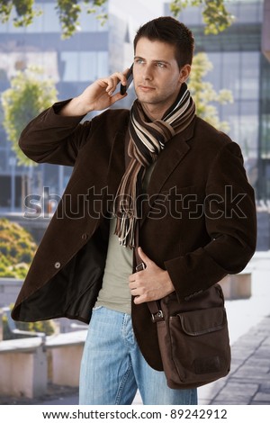 Goodlooking man on mobile phone call standing outside of office building in spring sun, wearing trendy bag and scarf.?