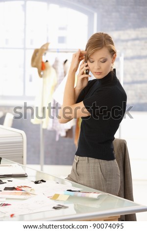 Young attractive female fashion designer talking on mobile phone in office, looking at patterns on desk.?