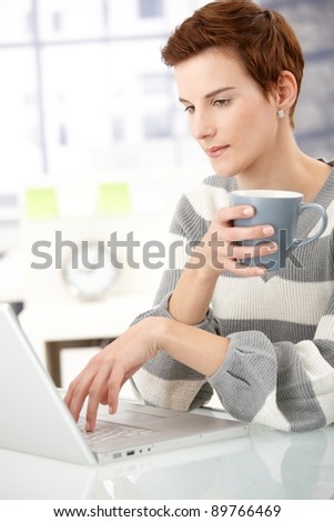 Office girl sitting at desk with mug handheld, working on laptop computer.?