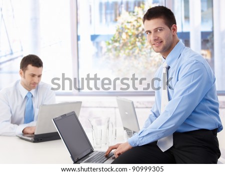Young businessman sitting on top of desk, working on laptop, smiling.?