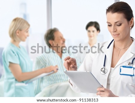 Doctor recording information to electronic tablet at patients bed team working in background. Doctor recording information to electronic tablet at patients bed team working in background.