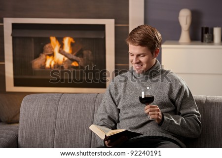 Young man sitting on sofa at home on a cold winter day, reading book in front of fireplace, tasting red wine.?