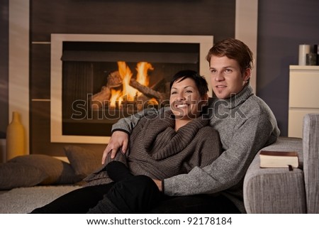 Young couple sitting on floor hugging in front of fireplace at home, looking away, smiling.?