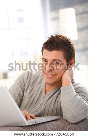 Portrait of young man using laptop computer at home, smiling at camera.?