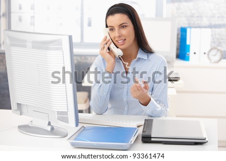 Attractive woman sitting at desk at work on landline phone call, gesturing, smiling.?