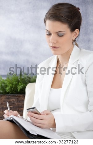 Businesswoman sitting in office using cellphone, writing notes into personal organizer.?