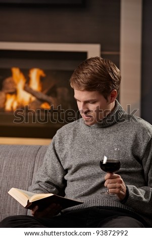 Young man sitting on sofa at home on a cold winter day, reading book in front of fireplace, tasting red wine.?