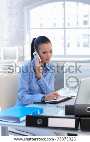 Businesswoman taking mobile phone call and writing notes into calendar at office desk.?