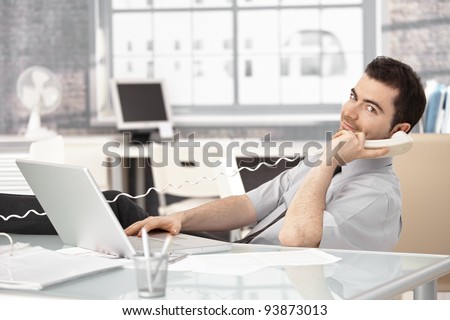 Young stock broker working in bright office, using laptop, talking on phone.?