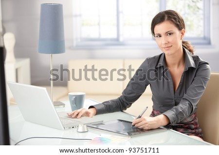 Attractive young graphic designer using laptop and tablet, working at home, smiling.?