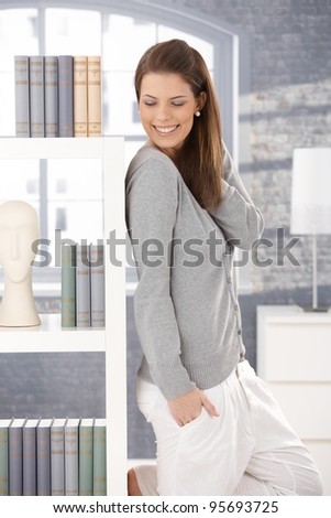 Happy woman wearing leisure clothes posing at book shelf at home, laughing.?
