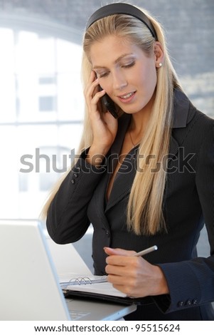 Smiling confident businesswoman writing notes into personal organizer, speaking on mobile phone, standing in office.?