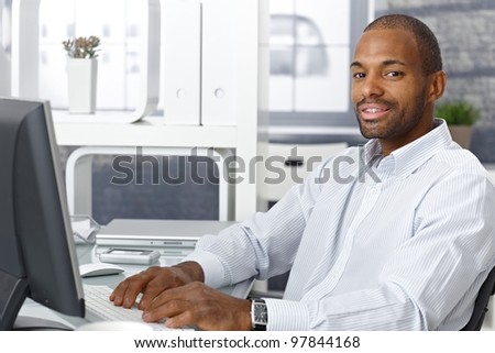 Businessman working on computer at office desk, looking at camera, smiling.