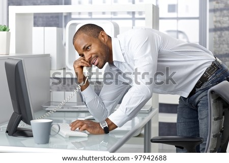 Casual happy businessman talking on landline phone in office, standing leaning on desk.