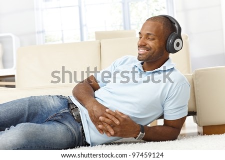 Handsome black man enjoying listening to music on headphones, lying on living room floor, smiling with eyes closed.