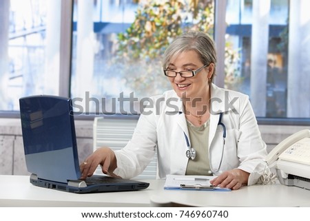 Happy female doctor working at desk in doctors room, using laptop, doing paperwork.