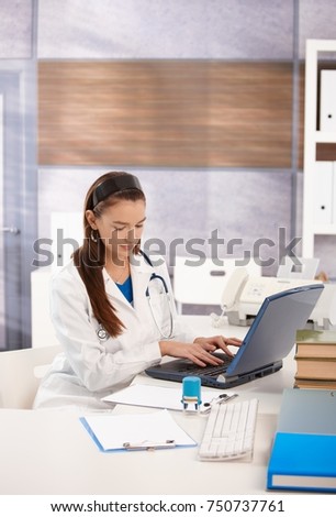 Young female doctor sitting at desk in doctors office using computer, working.