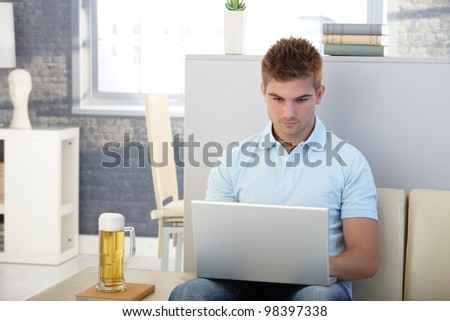 Young man sitting in living room using laptop computer, having beer.