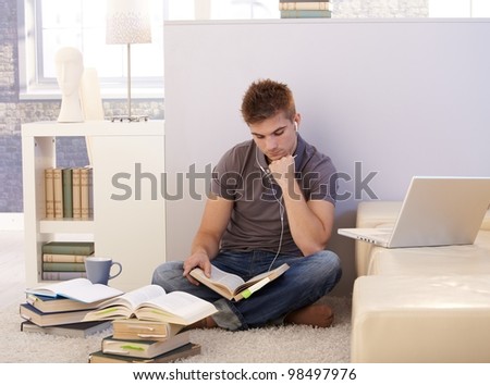 College student boy studying at home surrounded by books, notes, listening to music via headphones, sitting on living room floor.
