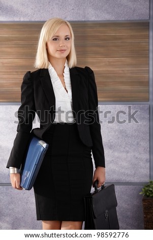 Portrait of smiling young businesswoman standing in office with laptop computer and briefcase handheld.