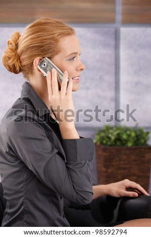 Businesswoman sitting in office lobby on mobile phone call, smiling, side view portrait.