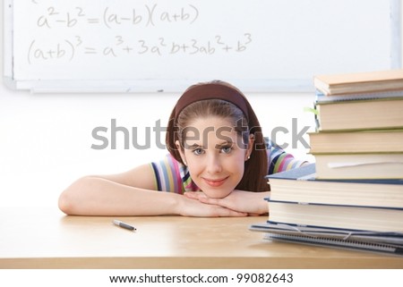 Teenage girl sitting at desk in classroom front of whiteboard, smiling at camera.