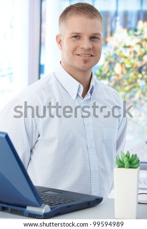 Office worker guy standing by laptop computer, smiling at camera.