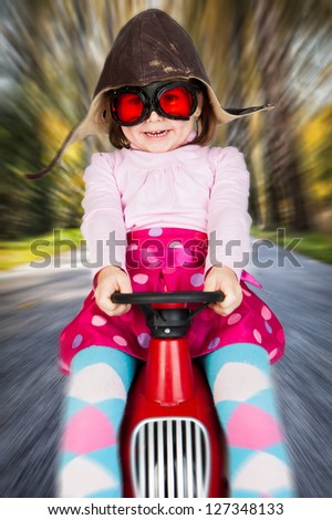 Girl in retro racing hat and goggles driving on toy car at speed with blurred background.