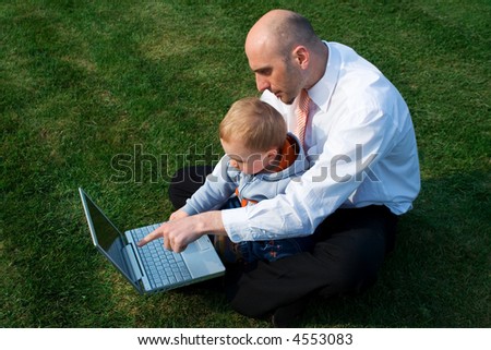 A father sits with his son as they use a laptop computer. The father is pointing to something on the screen. They are sitting outside on the green grass.