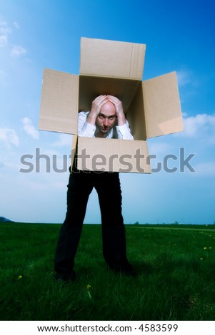 Man stands holding his hands on top of his head.  His torso is covered by a cardboard box and he is looking out from the top of the open box.  The background is green grass and blue sky.