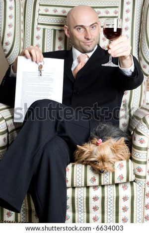 A successful realtor toasting/celebrating to a newly won deal, with a terrier at his side and a glass of red wine in hand, dons a black suit and sits on an upholstered armchair.