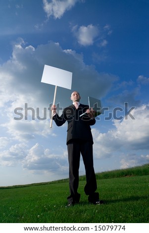 Bold man standing on the grass. He hold blank sign in one hand and laptop in other. Available space for text.